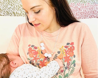 May include: A woman wearing a pink floral print sweatshirt with a white necklace featuring wooden and silicone beads. She is holding a baby wearing a white and gray floral print onesie.