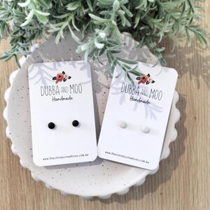 May include: Two pairs of stud earrings on white display cards labelled Dubba and Moo Handmade. One pair features small matte black circular studs, while the other pair features small matte white circular studs. The cards include the website address.