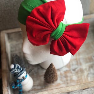 Puede incluir: Una diadema festiva con base verde y un gran lazo rojo. El lazo est&aacute; acentuado con una banda verde en el centro. La diadema se muestra en una cabeza de maniqu&iacute; blanca, un accesorio de moda.