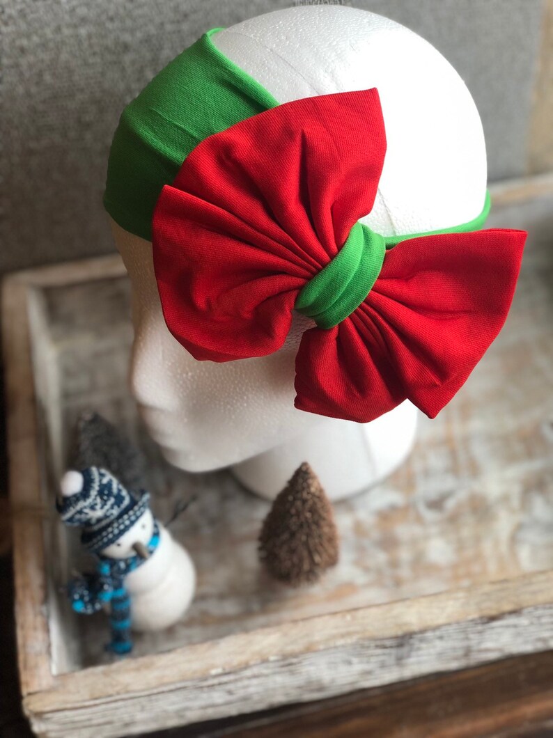 Puede incluir: Una diadema festiva con base verde y un gran lazo rojo. El lazo est&aacute; acentuado con una banda verde en el centro. La diadema se muestra en una cabeza de maniqu&iacute; blanca, un accesorio de moda.