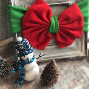 Puede incluir: Imagen con tem&aacute;tica navide&ntilde;a que presenta un lazo rojo y verde, una figura de mu&ntilde;eco de nieve con gorro y bufanda azules y blancos, y peque&ntilde;os &aacute;rboles decorativos. El lazo es el punto focal, sobre un fondo r&uacute;stico enmarcado.