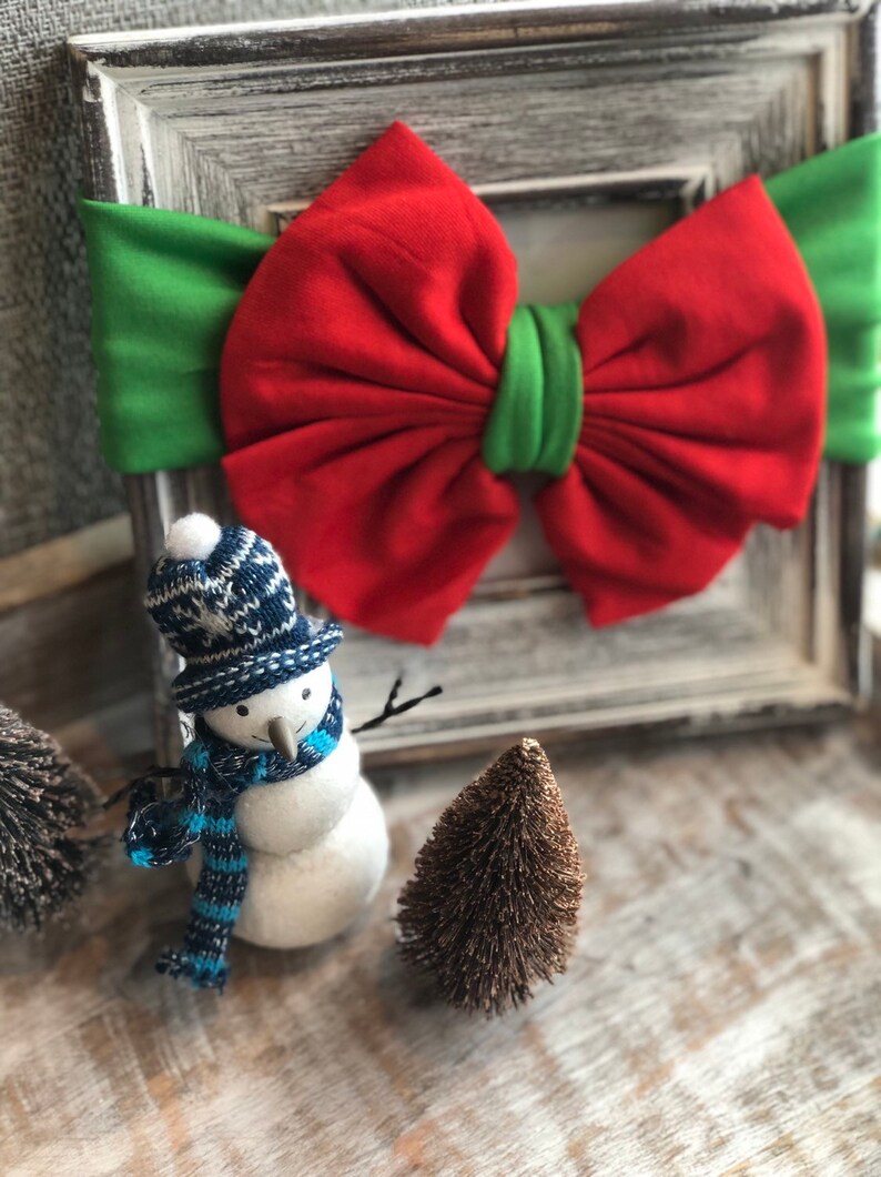 Puede incluir: Imagen con tem&aacute;tica navide&ntilde;a que presenta un lazo rojo y verde, una figura de mu&ntilde;eco de nieve con gorro y bufanda azules y blancos, y peque&ntilde;os &aacute;rboles decorativos. El lazo es el punto focal, sobre un fondo r&uacute;stico enmarcado.