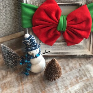 Puede incluir: Una diadema navide&ntilde;a festiva con un gran lazo rojo y detalles verdes. Un peque&ntilde;o mu&ntilde;eco de nieve con gorro y bufanda de punto azul y blanco est&aacute; junto a dos &aacute;rboles de Navidad en miniatura. Los art&iacute;culos est&aacute;n dispuestos sobre una superficie de madera r&uacute;stica.