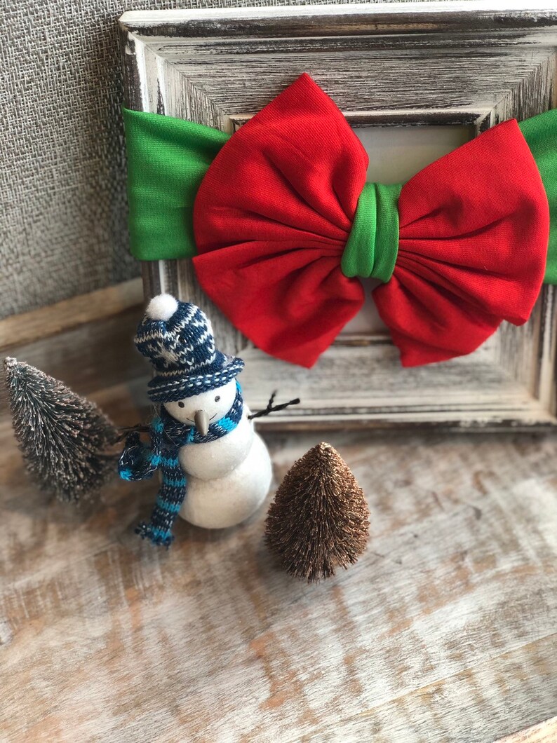 Puede incluir: Una diadema navide&ntilde;a festiva con un gran lazo rojo y detalles verdes. Un peque&ntilde;o mu&ntilde;eco de nieve con gorro y bufanda de punto azul y blanco est&aacute; junto a dos &aacute;rboles de Navidad en miniatura. Los art&iacute;culos est&aacute;n dispuestos sobre una superficie de madera r&uacute;stica.