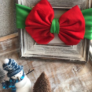 Puede incluir: Una diadema con lazo rojo y verde, con un lazo rojo y una banda verde, se encuentra dentro de un marco blanco. Un peque&ntilde;o mu&ntilde;eco de nieve con gorro y bufanda azules y blancos est&aacute; junto a un peque&ntilde;o &aacute;rbol marr&oacute;n. Tema navide&ntilde;o.