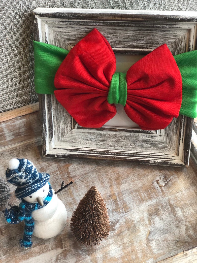 Puede incluir: Una diadema con lazo rojo y verde, con un lazo rojo y una banda verde, se encuentra dentro de un marco blanco. Un peque&ntilde;o mu&ntilde;eco de nieve con gorro y bufanda azules y blancos est&aacute; junto a un peque&ntilde;o &aacute;rbol marr&oacute;n. Tema navide&ntilde;o.