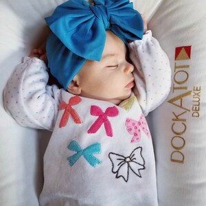 Puede incluir: Un beb&eacute; durmiendo con una diadema de lazo azul brillante. El beb&eacute; viste una camiseta blanca de manga larga adornada con aplicaciones de lazo de colores. El beb&eacute; descansa sobre una superficie acolchada blanca.