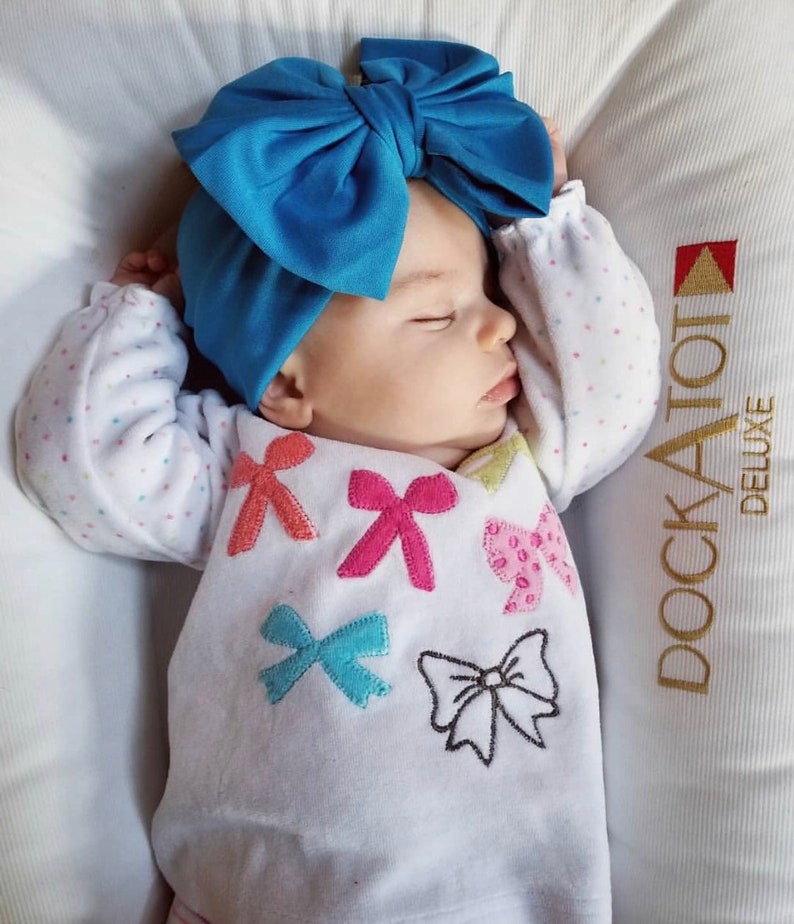 Puede incluir: Un beb&eacute; durmiendo con una diadema de lazo azul brillante. El beb&eacute; viste una camiseta blanca de manga larga adornada con aplicaciones de lazo de colores. El beb&eacute; descansa sobre una superficie acolchada blanca.
