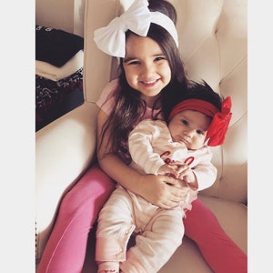 Puede incluir: Dos ni&ntilde;as peque&ntilde;as sentadas en una silla blanca acolchada. La mayor lleva una camiseta rosa, pantalones rosas y una diadema blanca con lazo. La m&aacute;s peque&ntilde;a lleva un body a rayas rosas y blancas con una diadema roja con lazo.