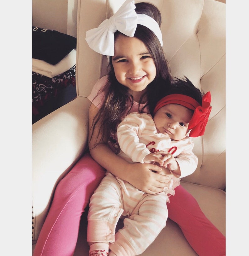 Puede incluir: Dos ni&ntilde;as peque&ntilde;as sentadas en una silla blanca acolchada. La mayor lleva una camiseta rosa, pantalones rosas y una diadema blanca con lazo. La m&aacute;s peque&ntilde;a lleva un body a rayas rosas y blancas con una diadema roja con lazo.