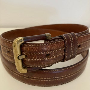 May include: A brown leather belt with a gold buckle and double stitching. The belt is folded in half.