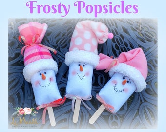 Puede incluir: Tres paletas de helado de muñeco de nieve con sombreros rosas y blancos. Las paletas de helado de muñeco de nieve están sobre una superficie con estampado blanco y negro. El texto "Frosty Popsicles Tutorial" está en la parte inferior de la imagen.