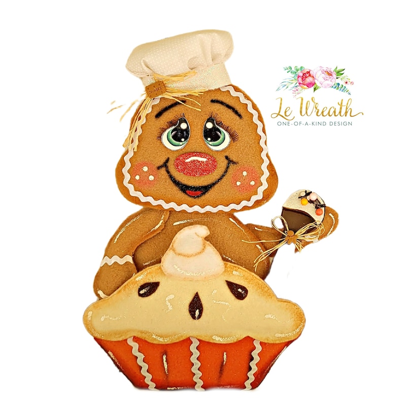 Puede incluir: Un hombre de jengibre con un gorro de cocinero sostiene un peque&ntilde;o cupcake y se sienta encima de un pastel con una cobertura de crema batida.  El hombre de jengibre tiene una cara sonriente con ojos grandes y mejillas sonrosadas.  El pastel est&aacute; decorado con una corteza marr&oacute;n y un relleno blanco.  El texto "Le Wreath One-of-a-Kind Design" es visible en la esquina superior derecha de la imagen.