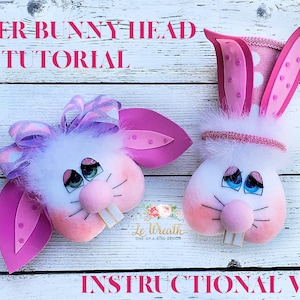 Könnte beinhalten: Zwei Osterhasenköpfe, einer mit einer rosa Schleife und der andere mit einem rosa gepunkteten Hut, beide mit weißem Fell, rosa Nasen und bemalten Augen. Der Text "Easter Bunny Head Tutorial" und "Instructional Video" ist auf dem weißen Holzgrund sichtbar.