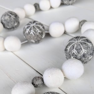 May include: A white and gray felt ball garland with snowflake designs. The garland is strung together with white thread and is perfect for decorating for the winter holidays.