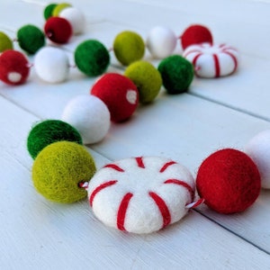 May include: A festive garland featuring felt balls and peppermint candies. The garland includes red, green, white, and lime green felt balls, along with white peppermint candies with red stripes. The garland is strung together with a white string.