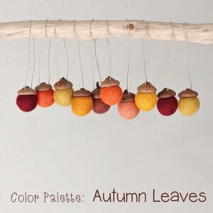 May include: A collection of felt acorn ornaments in shades of red, orange, and yellow, hanging from a branch. The acorns are arranged in a gradient pattern, with the darkest red at one end and the lightest yellow at the other. The text "Color Palette: Autumn Leaves" is written below the acorns.