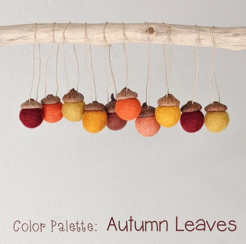 May include: A collection of felt acorn ornaments in shades of red, orange, and yellow, hanging from a branch. The acorns are arranged in a gradient pattern, with the darkest red at one end and the lightest yellow at the other. The text "Color Palette: Autumn Leaves" is written below the acorns.