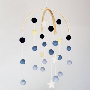 May include: A wooden baby mobile with blue, black, and yellow felt balls and stars. The mobile is suspended from a wooden hoop.