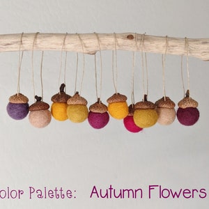 May include: A string of felt acorn ornaments in shades of purple, pink, yellow, and brown. The acorns are hanging from a branch and the text "Color Palette: Autumn Flowers" is below the acorns.