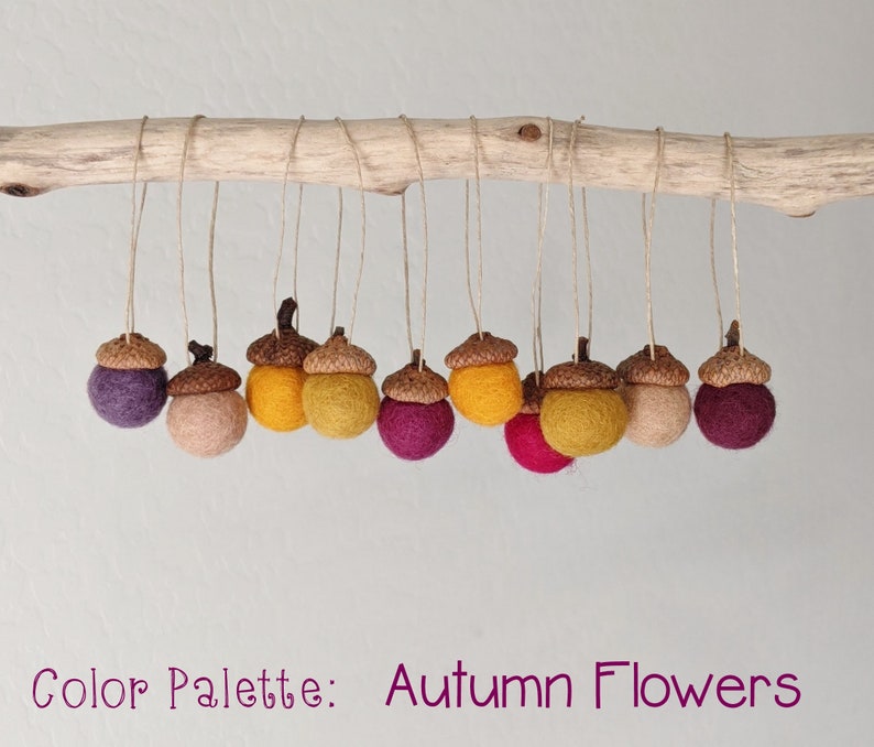 May include: A string of felt acorn ornaments in shades of purple, pink, yellow, and brown. The acorns are hanging from a branch and the text "Color Palette: Autumn Flowers" is below the acorns.