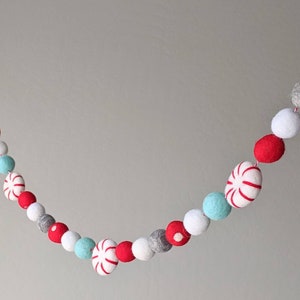 May include: A festive garland featuring red, white, gray, and light blue felt balls and peppermint candies strung on a red and white twine. The garland is a decorative item for the holidays.