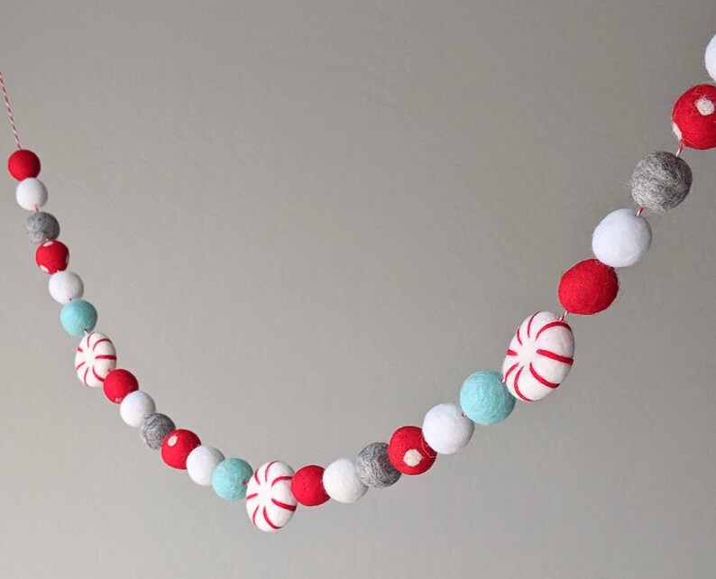 May include: A festive garland featuring red, white, gray, and light blue felt balls and peppermint candies strung on a red and white twine. The garland is a decorative item for the holidays.