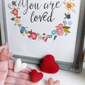 May include: A felt garland with red, pink, and white hearts strung together. The garland is hanging on a hand and in front of a framed print that says "You are loved" with a floral wreath around the text.