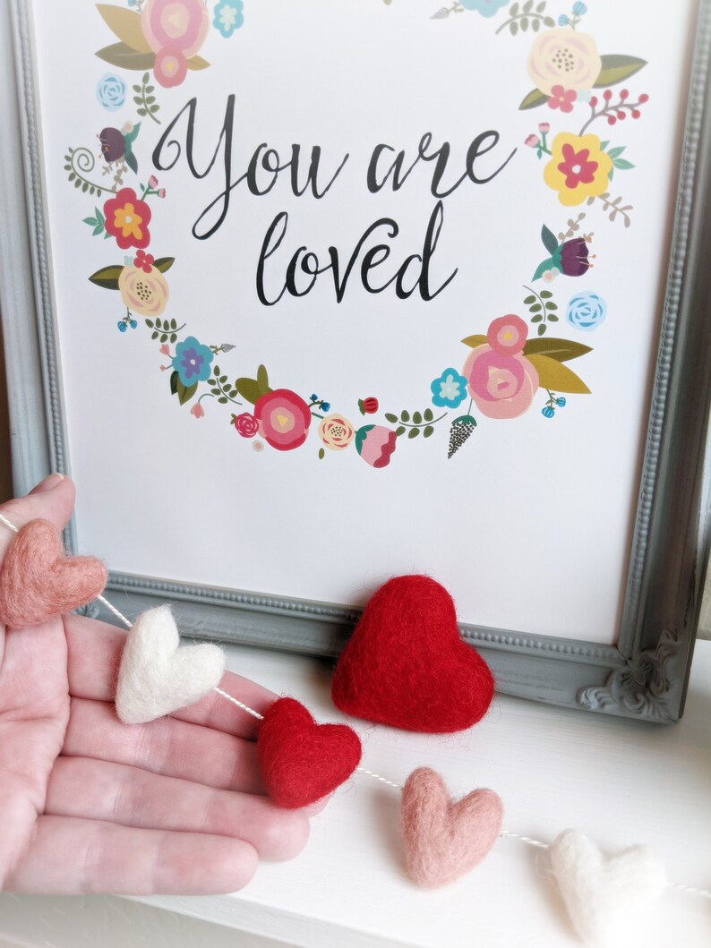 May include: A felt garland with red, pink, and white hearts strung together. The garland is hanging on a hand and in front of a framed print that says "You are loved" with a floral wreath around the text.