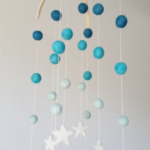 Op de afbeelding: Een babymobiel met een houten frame en blauwe en witte viltballen en sterren. De mobiel hangt aan het plafond.