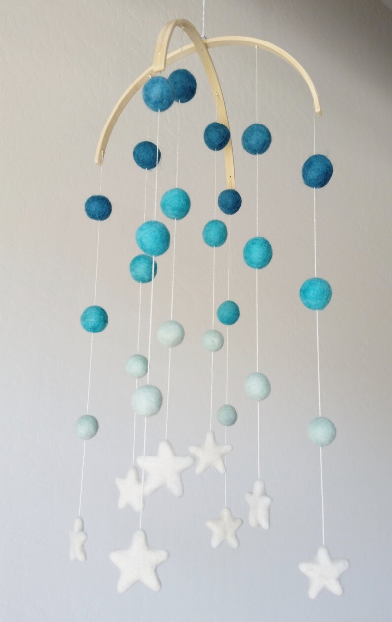 Op de afbeelding: Een babymobiel met een houten frame en blauwe en witte viltballen en sterren. De mobiel hangt aan het plafond.