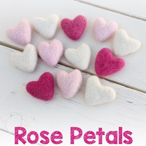 May include: A collection of felt hearts in three colors: fuchsia, pastel pink, and ivory. The hearts are arranged in a circle on a white wooden surface.