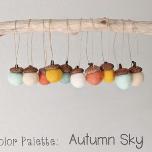 May include: A set of felt acorn ornaments in shades of brown, orange, yellow, and blue. The ornaments are hanging from a branch and the text "Color Palette: Autumn Sky" is written below.
