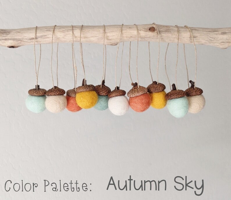 May include: A set of felt acorn ornaments in shades of brown, orange, yellow, and blue. The ornaments are hanging from a branch and the text "Color Palette: Autumn Sky" is written below.