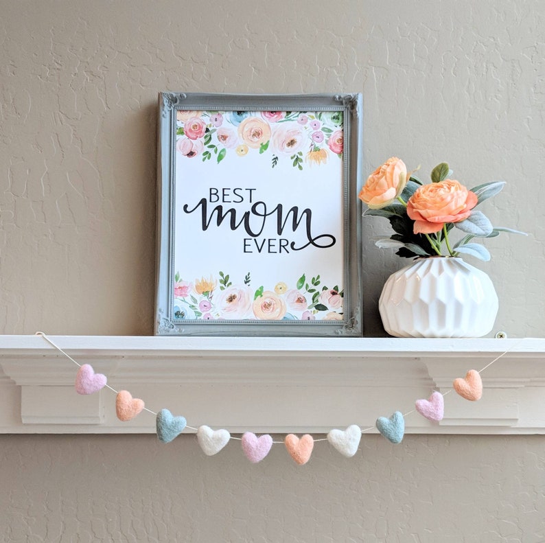 May include: A framed print with a floral design and the text "Best Mom Ever". A white vase with peach flowers sits on a white shelf with a garland of pastel felt hearts.