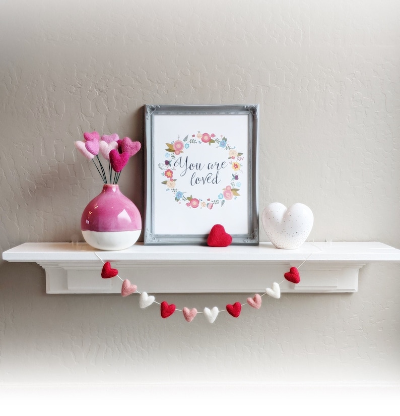 May include: A white shelf with a gray framed print that says "You are loved" surrounded by a floral wreath. A pink vase with pink felt hearts is on the left side of the shelf. A white felt heart is on the right side of the shelf. A white string of felt hearts hangs below the shelf.