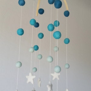Op de afbeelding: Een babymobiel met een houten frame en blauwe en witte viltballen en sterren. De mobiel hangt aan het plafond.