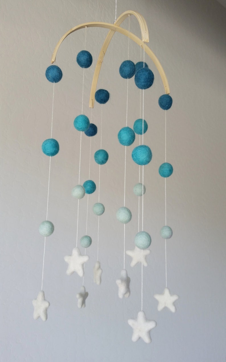 Op de afbeelding: Een babymobiel met een houten frame en blauwe en witte viltballen en sterren. De mobiel hangt aan het plafond.