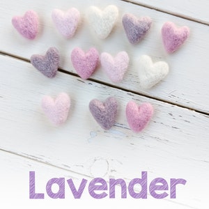 May include: A collection of felt hearts in shades of lavender, pink, and ivory. The hearts are arranged in a heart shape on a white wooden background. The word "Lavender" is written in purple text below the hearts.