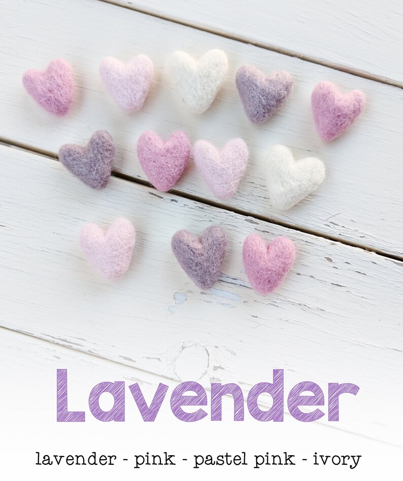 May include: A collection of felt hearts in shades of lavender, pink, and ivory. The hearts are arranged in a heart shape on a white wooden background. The word "Lavender" is written in purple text below the hearts.