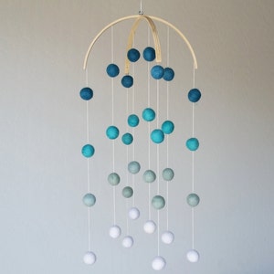 Op de afbeelding: Een houten mobiel met witte, lichtblauwe en donkerblauwe viltballen die aan een houten boog hangen. De ballen zijn gerangschikt in een verloop, met de witte ballen onderaan en de donkerblauwe ballen bovenaan.