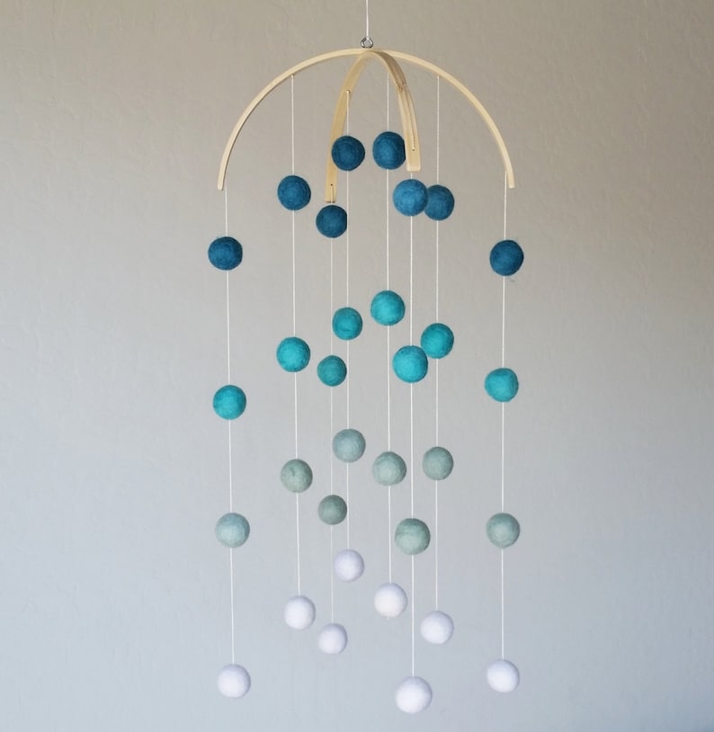 Op de afbeelding: Een houten mobiel met witte, lichtblauwe en donkerblauwe viltballen die aan een houten boog hangen. De ballen zijn gerangschikt in een verloop, met de witte ballen onderaan en de donkerblauwe ballen bovenaan.
