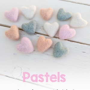 May include: A collection of felt heart shapes in pastel colors, including peach, seafoam, pastel pink, and ivory. The hearts are arranged in a heart shape on a white wooden surface. The text "Pastels" is written in pink below the hearts.