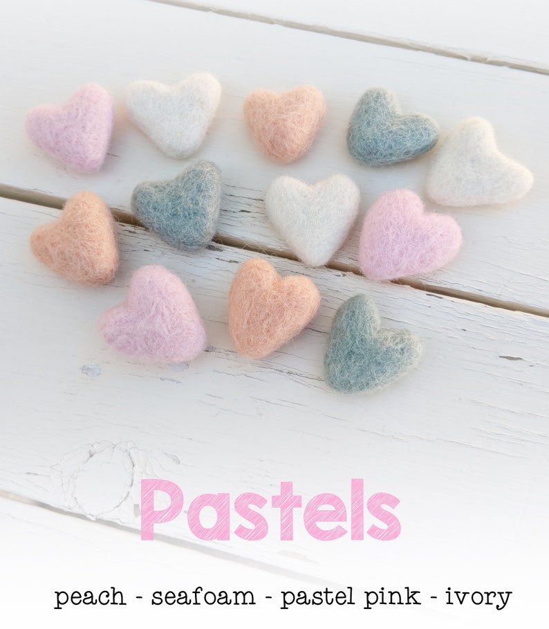May include: A collection of felt heart shapes in pastel colors, including peach, seafoam, pastel pink, and ivory. The hearts are arranged in a heart shape on a white wooden surface. The text "Pastels" is written in pink below the hearts.