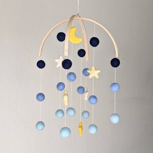 May include: A wooden baby mobile with a crescent moon and stars. The mobile features blue, yellow, and white felt balls and stars.
