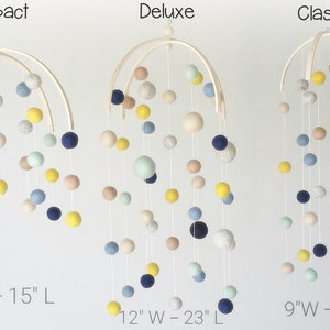 Op de afbeelding: Drie houten babymobielen met viltballen in verschillende kleuren, waaronder blauw, geel, beige en groen. De mobielen zijn gelabeld "Compact" met een breedte van 30 cm en een lengte van 38 cm, "Deluxe" met een breedte van 30 cm en een lengte van 58 cm, en "Classic" met een breedte van 23 cm en een lengte van 51 cm.