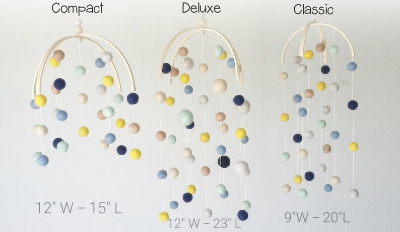 Op de afbeelding: Drie houten babymobielen met viltballen in verschillende kleuren, waaronder blauw, geel, beige en groen. De mobielen zijn gelabeld "Compact" met een breedte van 30 cm en een lengte van 38 cm, "Deluxe" met een breedte van 30 cm en een lengte van 58 cm, en "Classic" met een breedte van 23 cm en een lengte van 51 cm.