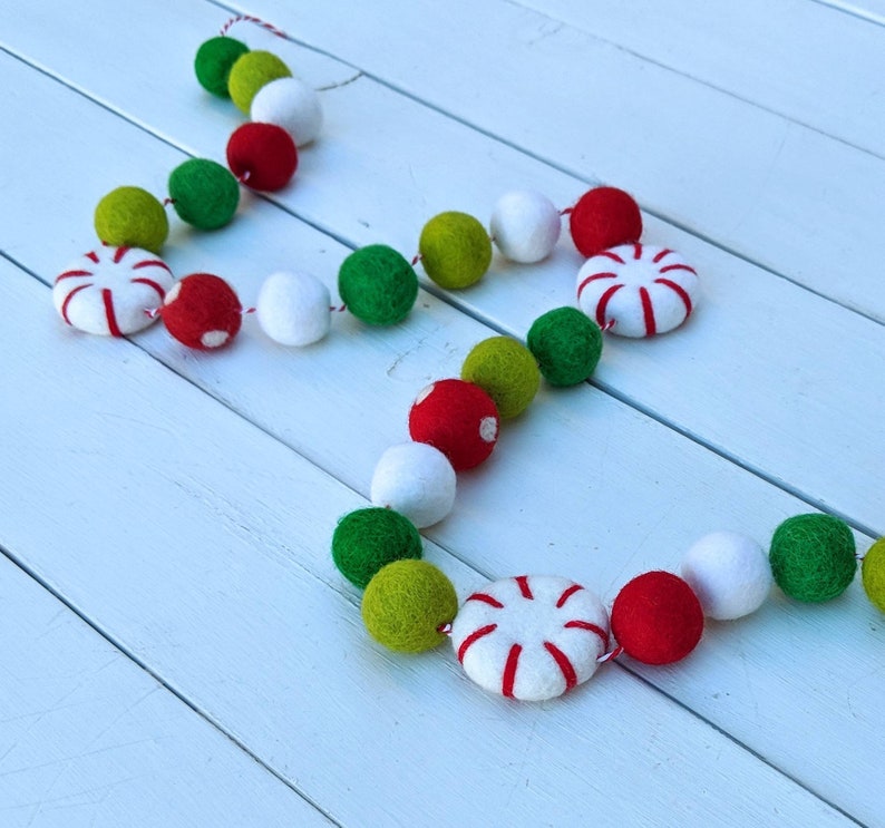 May include: A festive garland featuring red, white, green, and yellow felt balls and peppermint-shaped ornaments. The garland is strung on a red and white twine, perfect for holiday decorations. The felt balls are approximately 1 inch in diameter.
