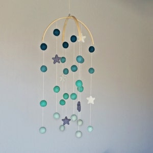 Op de afbeelding: Een houten mobiel met blauwe, groene en paarse viltballen en witte viltsterren die aan een houten ring hangen.
