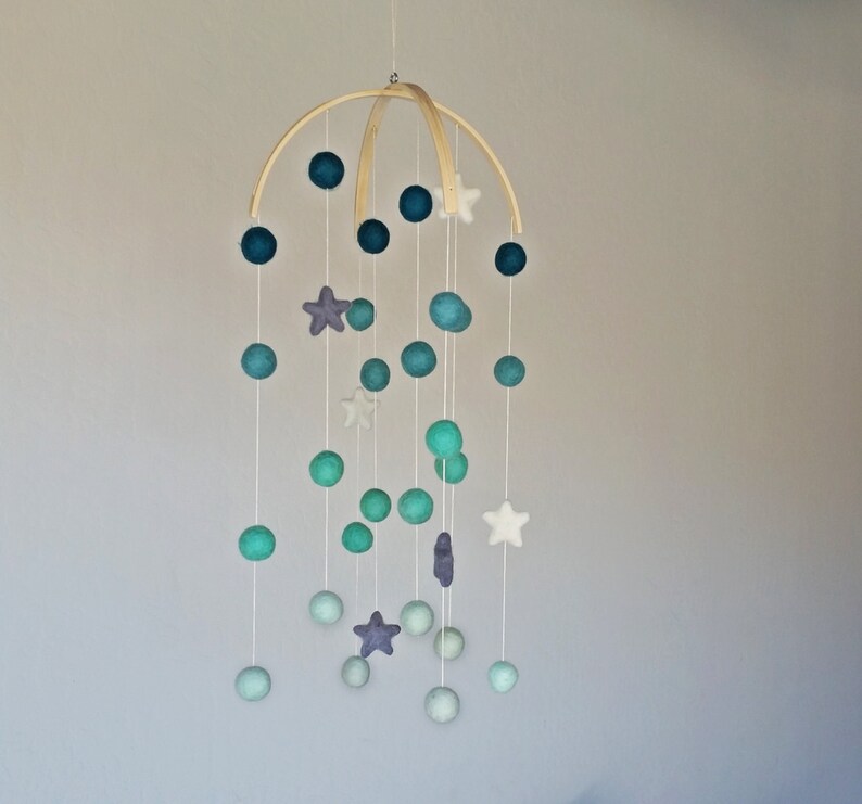 Op de afbeelding: Een houten mobiel met blauwe, groene en paarse viltballen en witte viltsterren die aan een houten ring hangen.